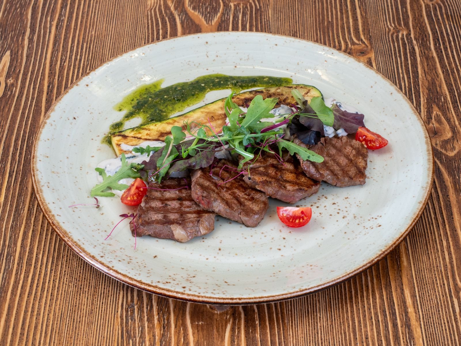 Grilled beef slices with a grilled zucchini strip, cherry tomatoes and leafy greens on a speckled plate